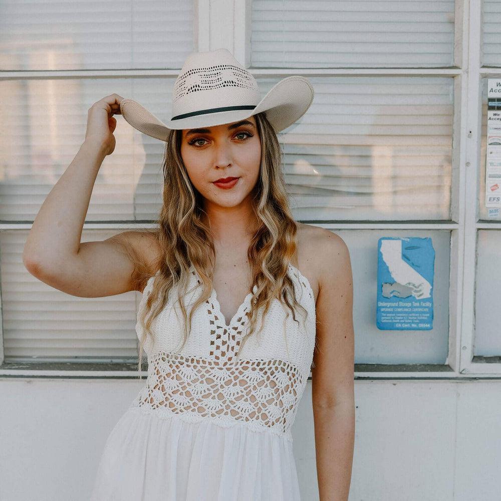 Woman wearing a white lace dress and cowboy hat in front of a building with a sign.